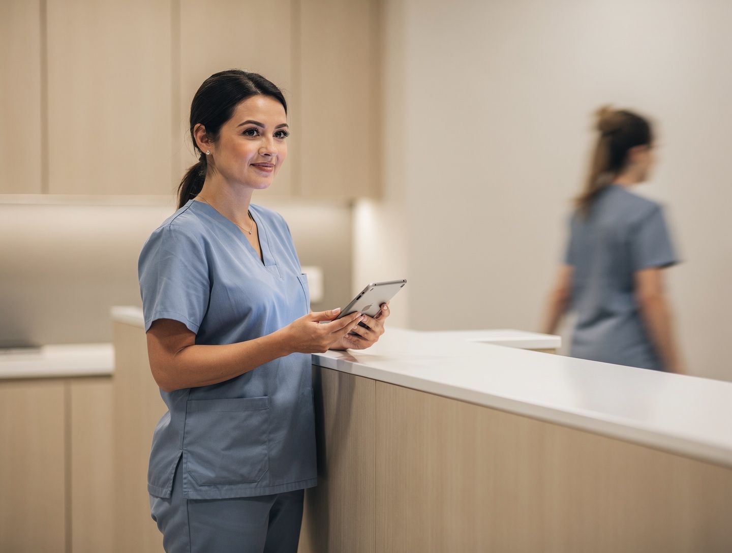 A dental assistant in blue scrubs holding a tablet at a clinic front desk.